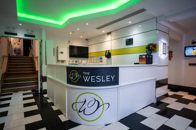 A reception desk with a The Wesley logo panel stands in a lobby under green ceiling lighting, with a staircase on the left and a black and white checkerboard floor.