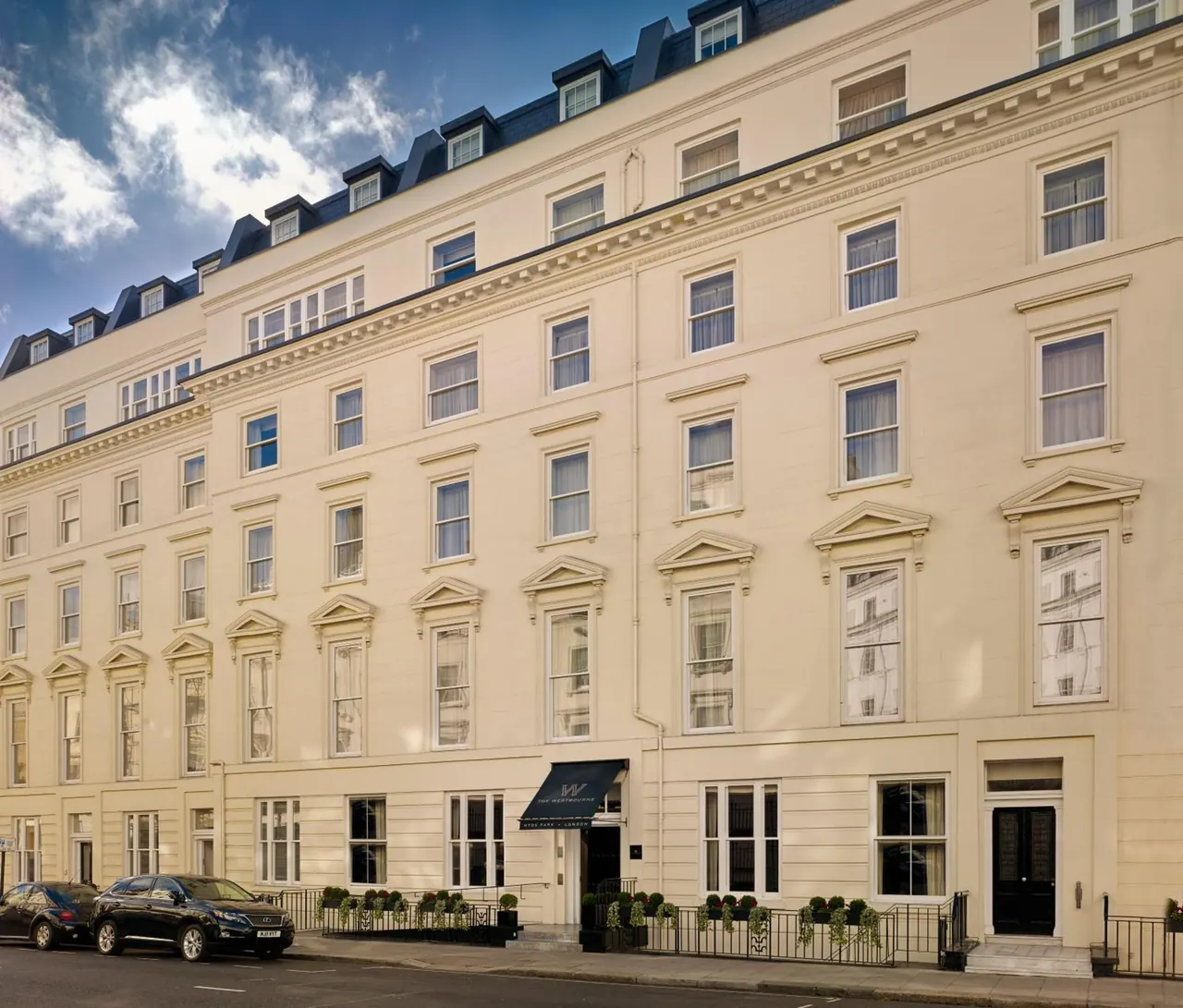 A cream stucco facade of a multi-storey hotel with a black awning over the entrance, with two dark cars parked on the street in front.