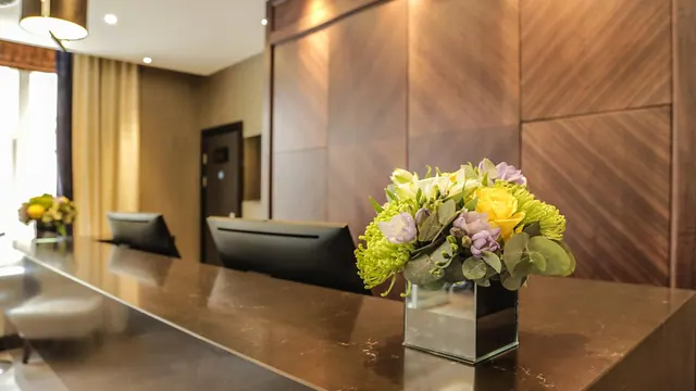 A reception desk with a flower arrangement in a mirrored glass vase, two monitors on the counter, and a window with curtains in the background.
