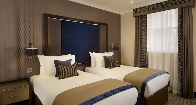 A twin bedroom with two single beds, a navy tufted headboard, wall lamps and a window with sheer curtains.
