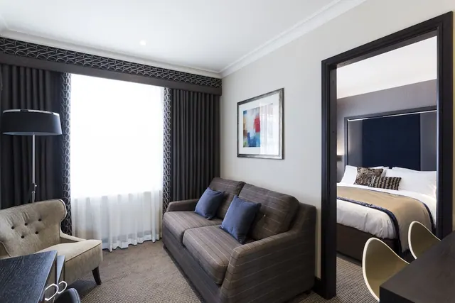 A living area with a striped grey sofa, blue cushions, an armchair and a floor lamp, with a bed visible through the open doorway to the bedroom.