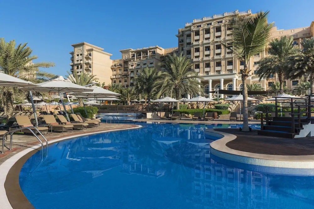 A large pool with sun loungers and shade umbrellas sits in front of a palm-lined hotel building under a clear blue sky.
