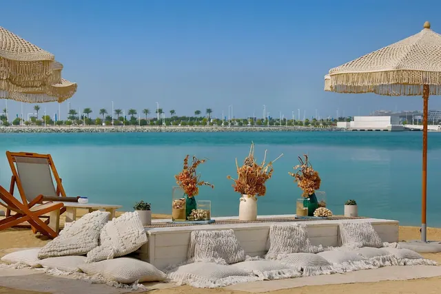 A staged beach seating area with macrame parasols, white floor cushions, dried flower vases and a view across turquoise water to palm trees and buildings.