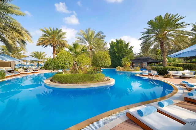 A large curved swimming pool with a planted island, palm trees, and sun loungers with blue towels in the foreground.