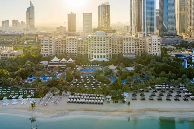 Aerial view of a beachfront hotel complex with several pools, rows of loungers and umbrellas on the sand, and the sea and city towers beyond.