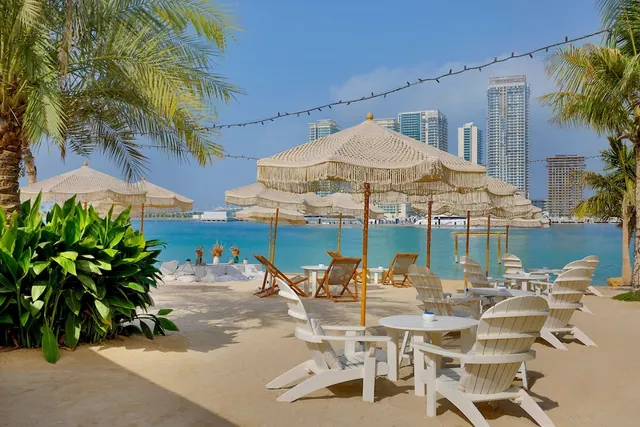 White Adirondack chairs and round tables sit under fringed beige umbrellas on a sandy beach, with turquoise water and high-rise towers beyond.