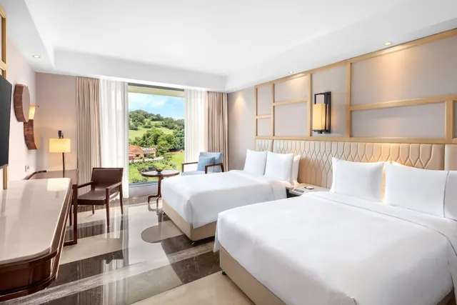 A bright hotel bedroom contains two large beds, a desk, seating, and a clear view of green hills through the window.