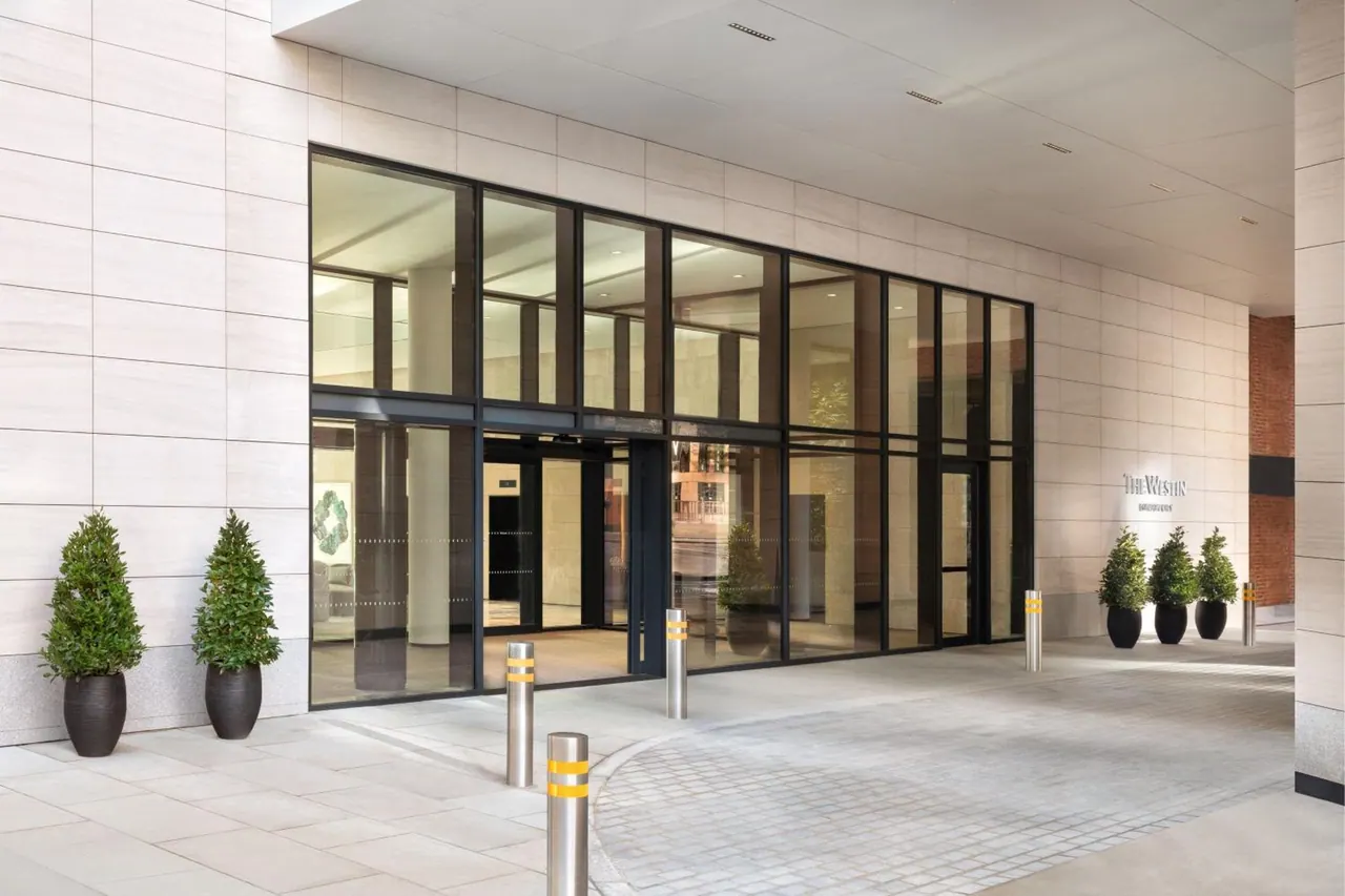 The glass-fronted entrance of The Westin hotel with steel bollards, potted shrubs and a wall sign, with no people visible.