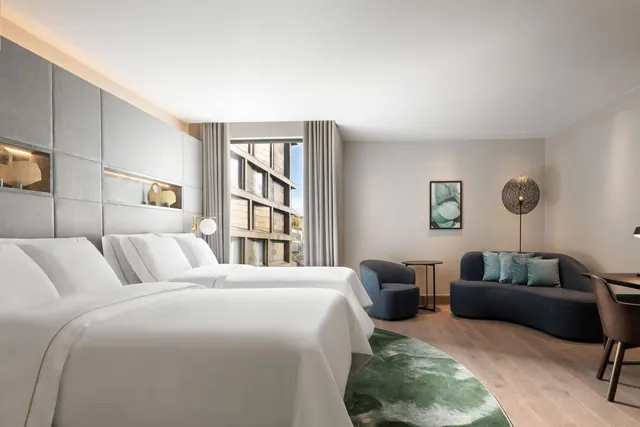 A hotel bedroom with two beds made up with white linen, a curved grey sofa and armchair, and a window looking onto a neighbouring building.