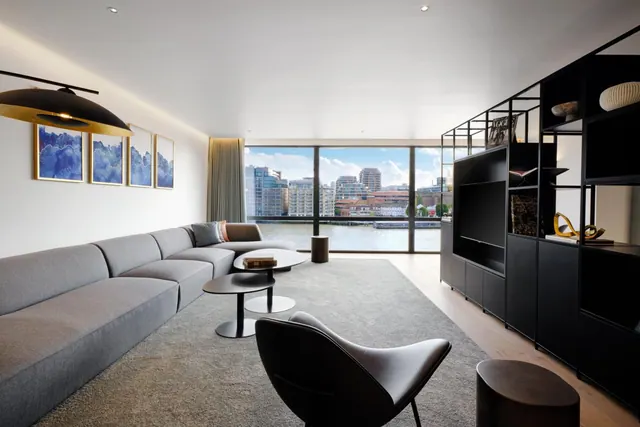 A living room with a grey sectional sofa, a black media unit with a wall-mounted television, and floor-to-ceiling windows looking over a river toward city buildings.
