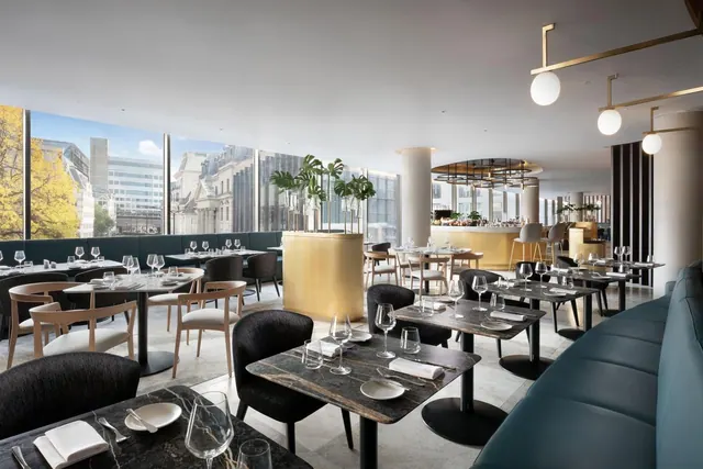 A restaurant dining room with marble-topped tables, teal banquette seating and tall windows looking out onto city buildings.