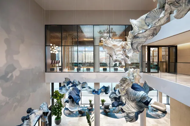A double-height lobby with large white and blue sculptural installations hanging from the ceiling and standing on the floor, with a glass-walled lounge and buildings visible through the windows beyond.