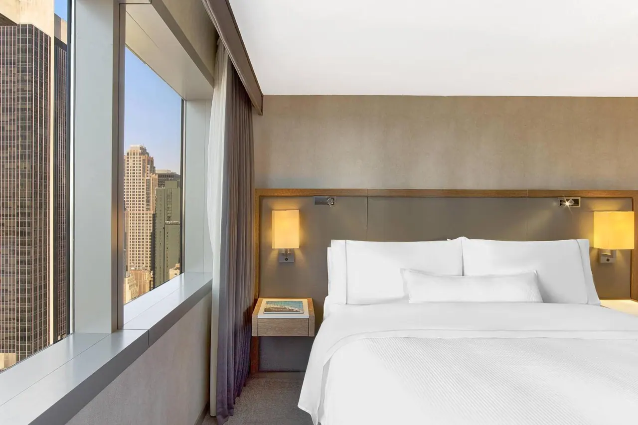 A made king bed with white linen sits beside a large window looking out over city high-rise buildings.