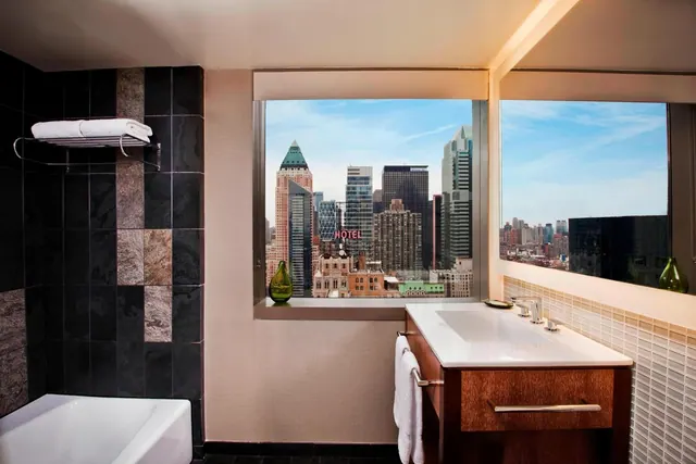 A bathroom with a white bathtub, a white vanity with a single sink, and large windows looking out over a city skyline with a hotel sign visible.