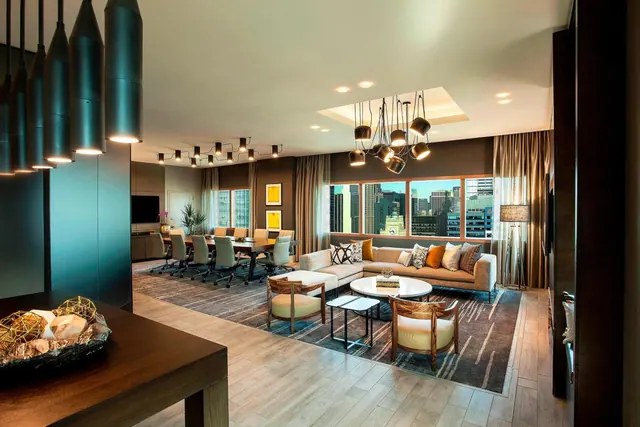 A living area with a beige sectional sofa, round coffee table and armchairs, with a conference table beyond and city buildings visible through the windows.