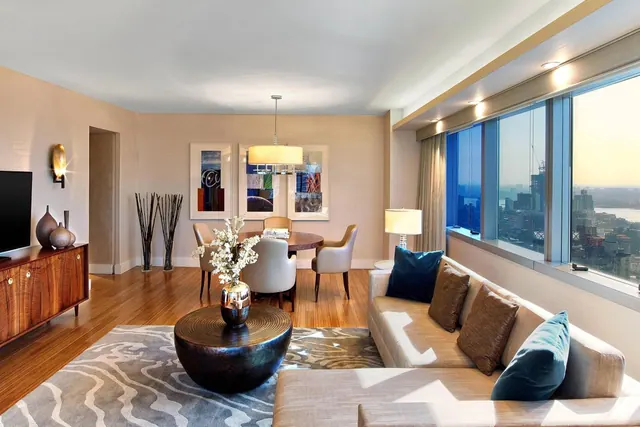A living and dining area with a sofa, a round dining table, a wall-mounted television and floor-to-ceiling windows overlooking a city skyline and a river.