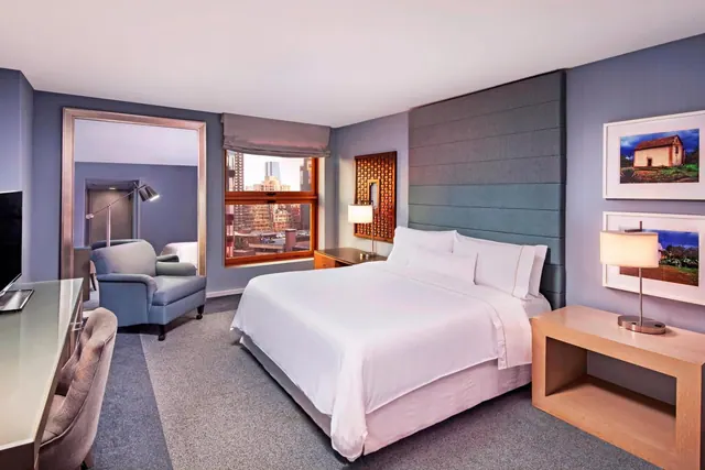 A hotel bedroom with a king bed, a grey armchair, a desk and television, and a window showing a city view.