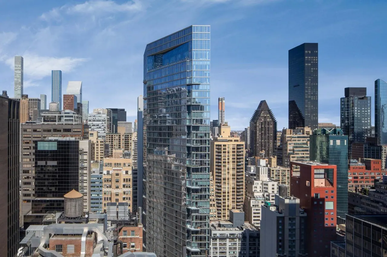 Elevated view of midtown Manhattan skyscrapers under a partly cloudy blue sky, with a rooftop water tank and low buildings in the foreground.