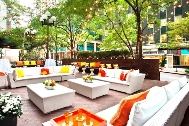 An outdoor lounge terrace with white wicker sofas, low tables, orange and yellow cushions, and string lights hanging over trees.