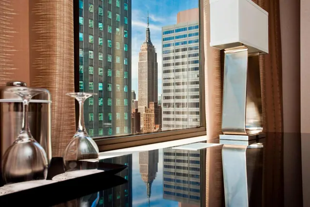A window in a room looks out at the Empire State Building among surrounding skyscrapers, with two wine glasses and a lamp on a dark table in the foreground.