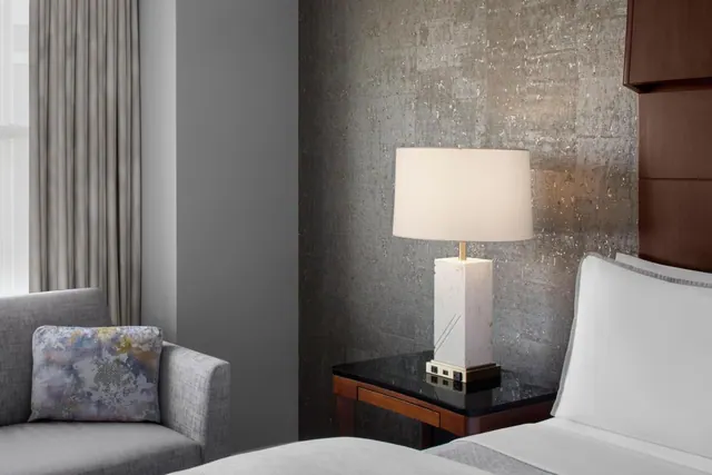 A bedroom with a grey armchair and patterned cushion, a white table lamp on a black-topped nightstand beside the bed, and a textured metallic wall.
