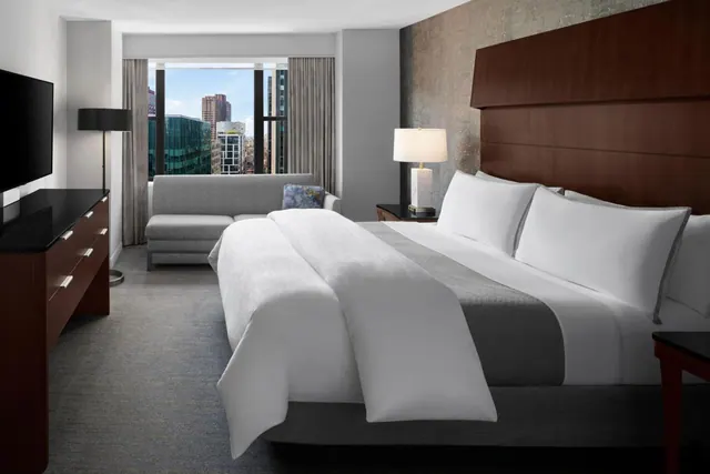 A king bed with white linens faces a large window with a city skyline view, beside a grey sofa and a wall-mounted television.
