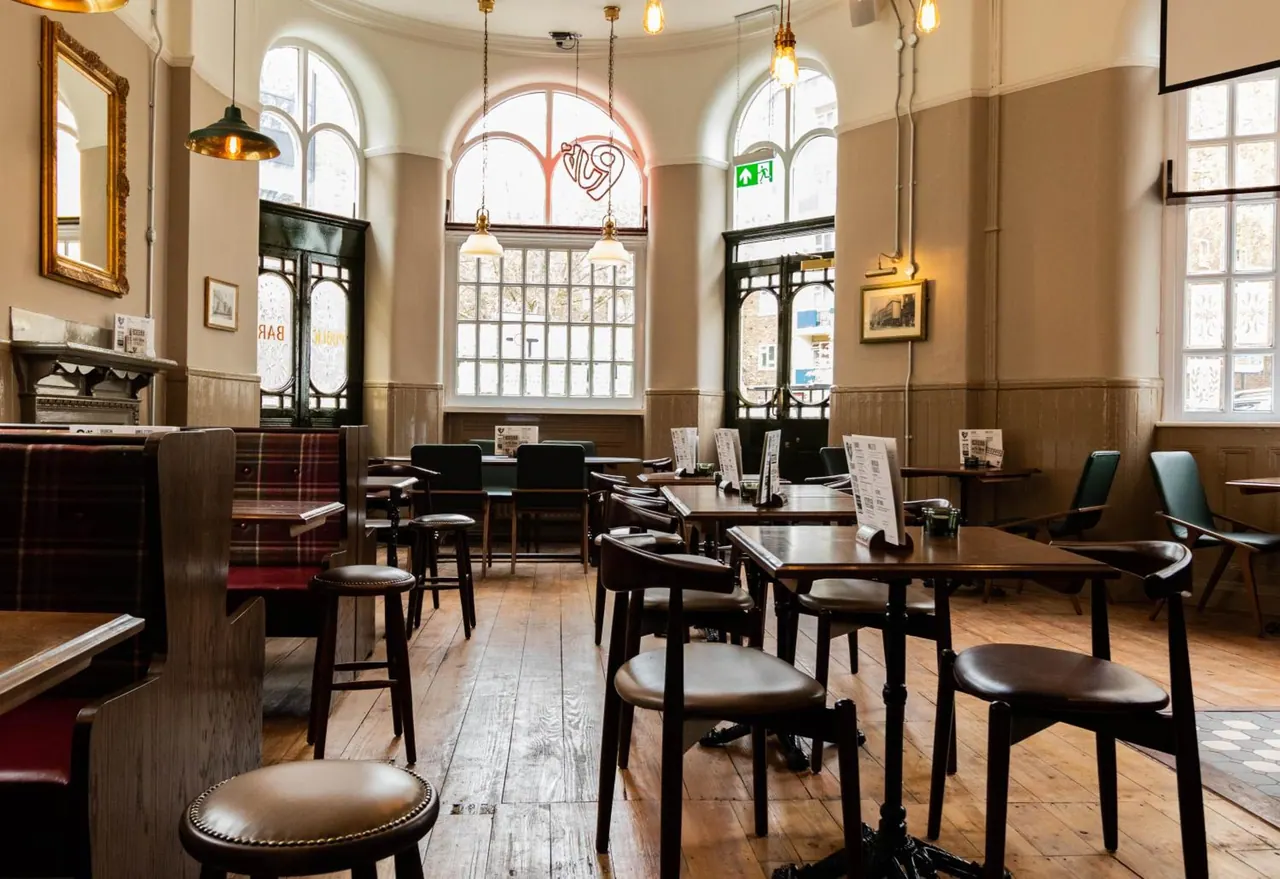 A dining room with wooden tables, leather-seated chairs, plaid booths, arched windows and pendant lights, with no people present.