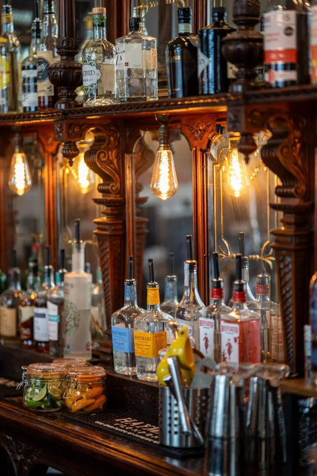 A bar counter with a carved wooden back bar holding bottles, with filament bulbs hanging above and jars of garnish on the counter.