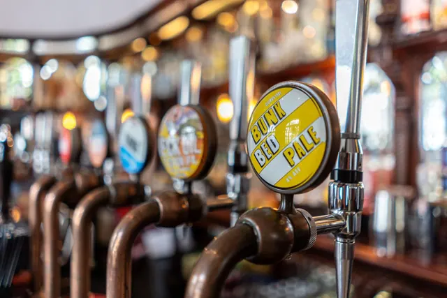 Close-up of a row of beer tap handles on a bar, with a yellow and white Bunk Bed Pale badge in the foreground and a blurred bar backdrop behind.