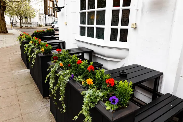 Black wooden planters with flowers and black tables line the pavement outside a white building with sash windows.