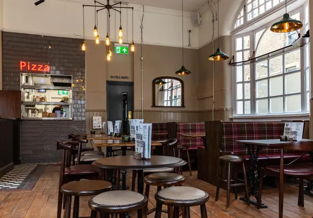 A dining room with wooden tables and stools, tartan banquette seating, pendant lights, and a pizza counter with a red neon sign.