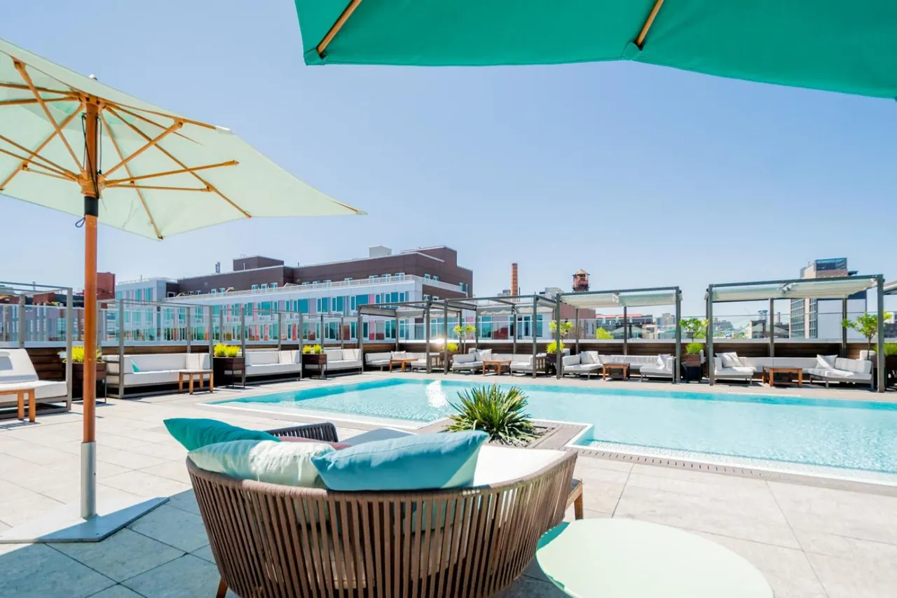 A rooftop pool with white lounge seating, wooden chairs, teal and cream umbrellas, and city buildings visible in the background.