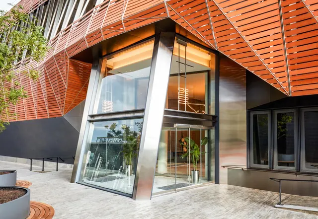 The entrance of a modern building with an orange slatted facade, angled glass walls and glass doors, with potted plants on the paved forecourt.