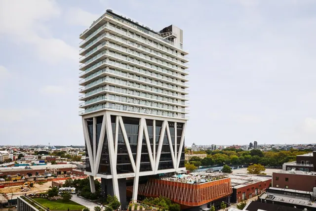 Exterior view of a tall hotel tower with stacked white balconies and angled white bracing at its base, with the name The William Vale on the lower facade and low buildings and trees around it.