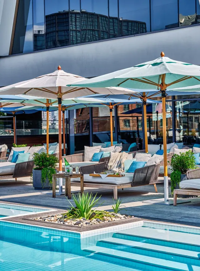 Pool deck with shaded umbrellas, cushioned lounge seating and planted greenery beside a turquoise pool with tiled steps, below a glass building facade.