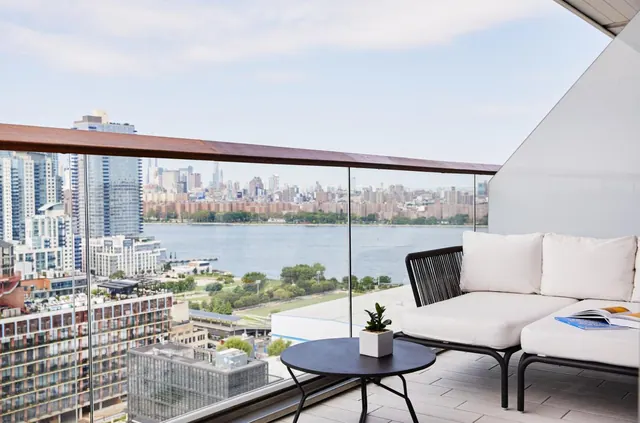 A private balcony with a white outdoor sofa, a round black side table and a potted plant, overlooking a river and a city skyline.