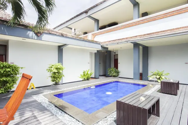A small rectangular blue-tiled pool sits in a courtyard surrounded by white-walled rooms, a wooden deck and pale stones.