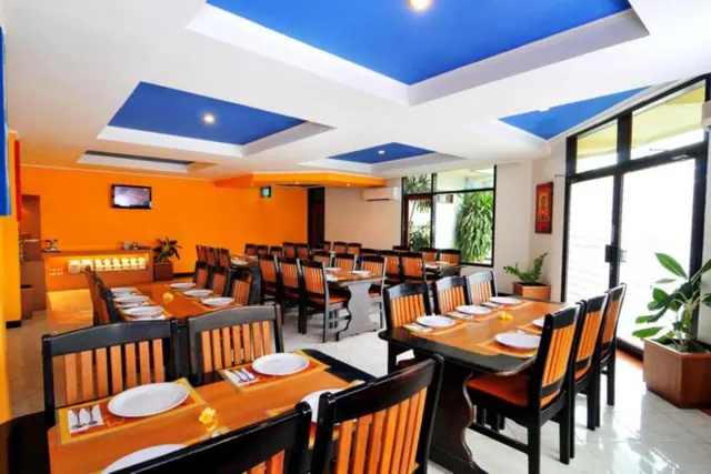 An empty dining room with orange walls, blue ceiling panels, wooden tables set with plates and napkins, and tall windows showing greenery outside.