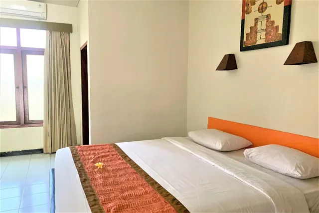 A bedroom with a double bed under an orange headboard, white pillows, a frangipani flower on the bed runner, wall art, wall sconces, and a wall-mounted air conditioner above a window with curtains.
