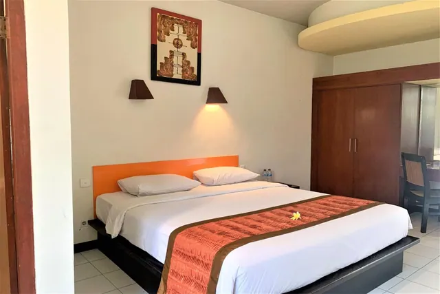 A bedroom with a made king bed under a wall painting and two wall lamps, next to a wooden wardrobe and a desk with a chair.
