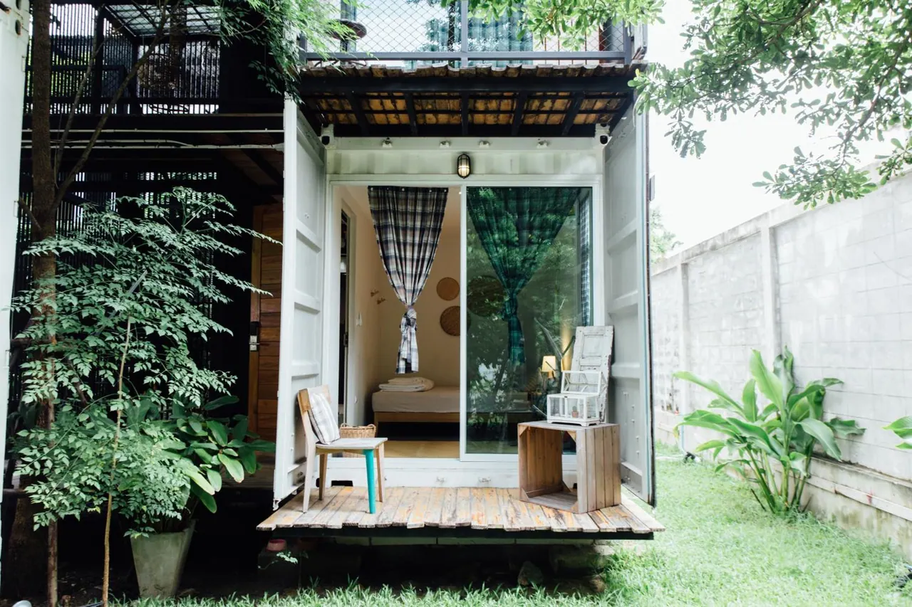 A converted shipping container room with open doors shows a wooden deck with a chair and a crate table, and a bed visible through the glass door, set in a garden.