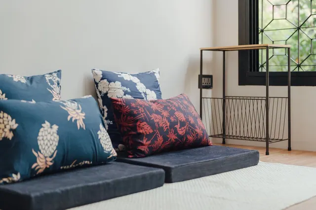 Patterned cushions sit on low floor mattresses beside a metal and wood side table and a window with a geometric grille showing greenery outside.