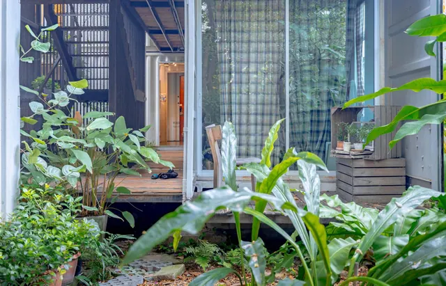 A wooden deck and glass sliding door of a container-style room, framed by potted plants and foliage in a garden courtyard.