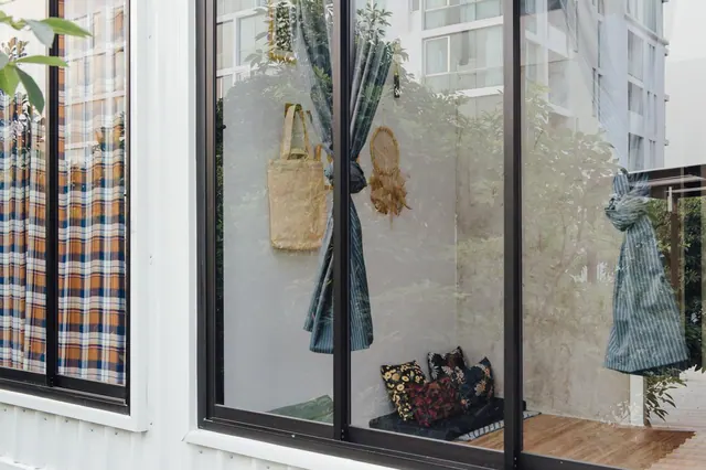 Exterior view through large glass windows into a room with plaid curtains, a hanging tote bag, a dreamcatcher and cushions on a low seat, with a wooden deck outside.