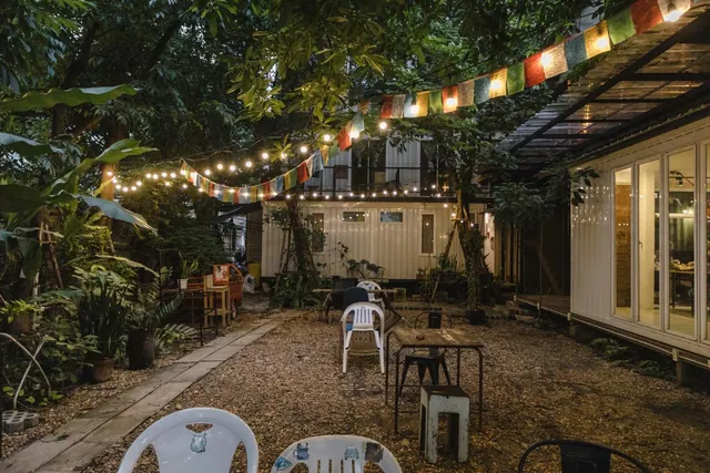 An evening courtyard garden with string lights, prayer flags, white plastic chairs and wooden tables on a gravel floor beside a glass-fronted building.