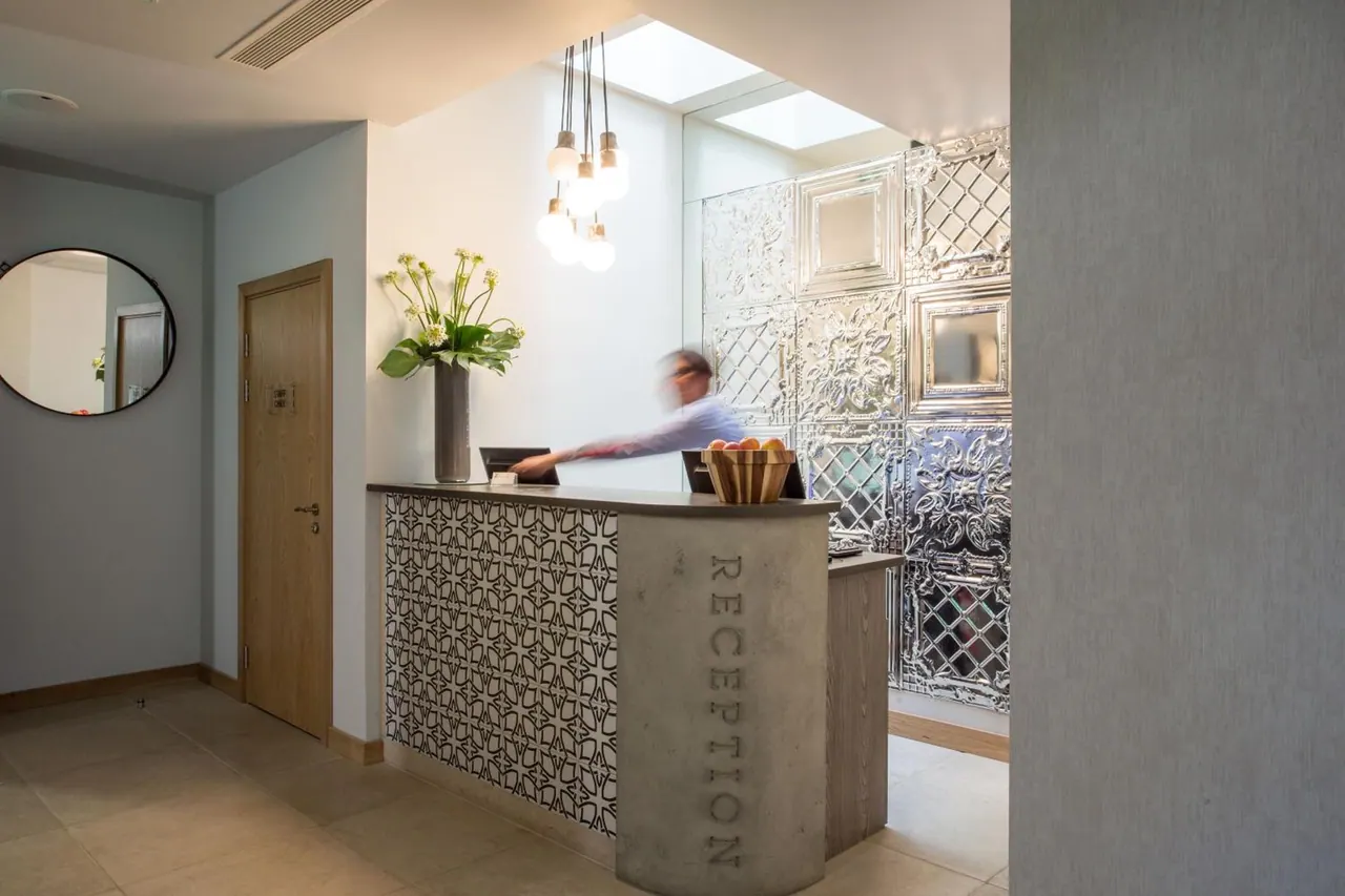 A blurred staff member stands behind a reception desk with a patterned tile front and a metal panel wall behind, under a row of pendant lights.