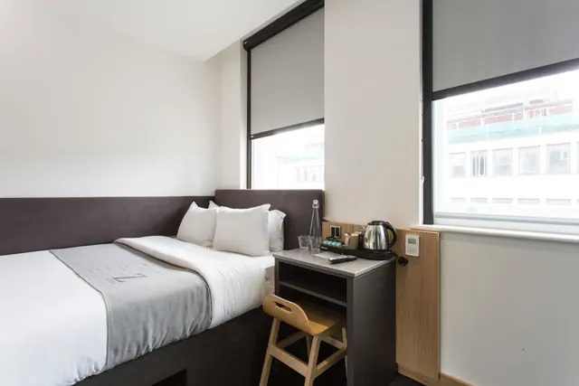A small hotel bedroom with a double bed, a bedside unit holding a kettle and glasses, a wooden stool, and two windows looking onto a neighbouring building.
