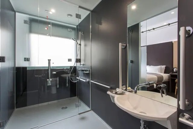 A modern bathroom with a glass-walled walk-in shower, grab rails, a wall-mounted basin, and a mirror reflecting a bed in the room beyond.