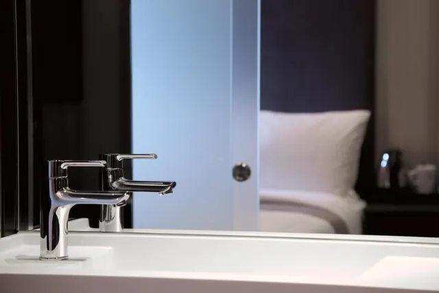 Chrome basin taps sit on a white counter in the foreground, with a bed and white pillows visible through a frosted glass door behind.