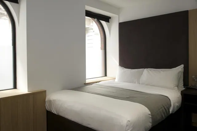 A double bed with a dark upholstered headboard sits beside two arched windows in a bedroom.